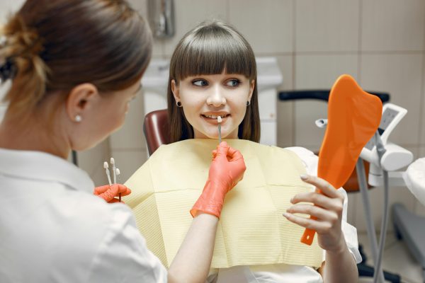 How Long Do Veneers Last? Lifespan of Porcelain vs. Composite cosmetic dentist holding a veneer sample up to a patient's tooth while the patient looks in a mirror